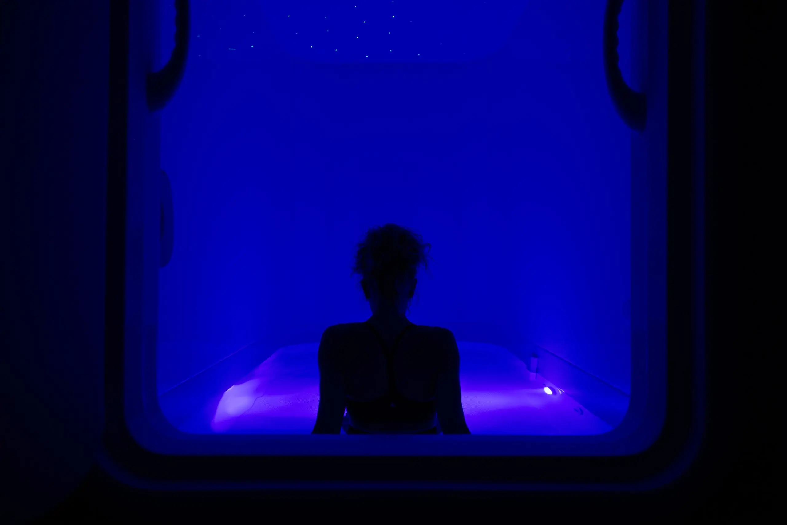 Guest sitting up in the float pod under blue light, seen through the open door